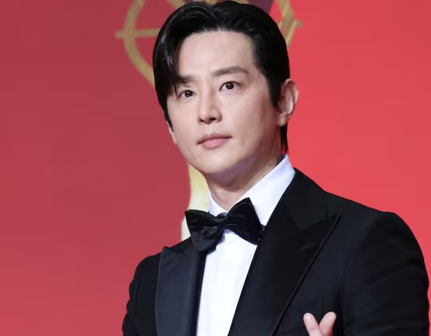 검은 턱시도와 나비넥타이를 착용한 남성이 붉은색 배경 앞에서 포즈를 취하고 있다.