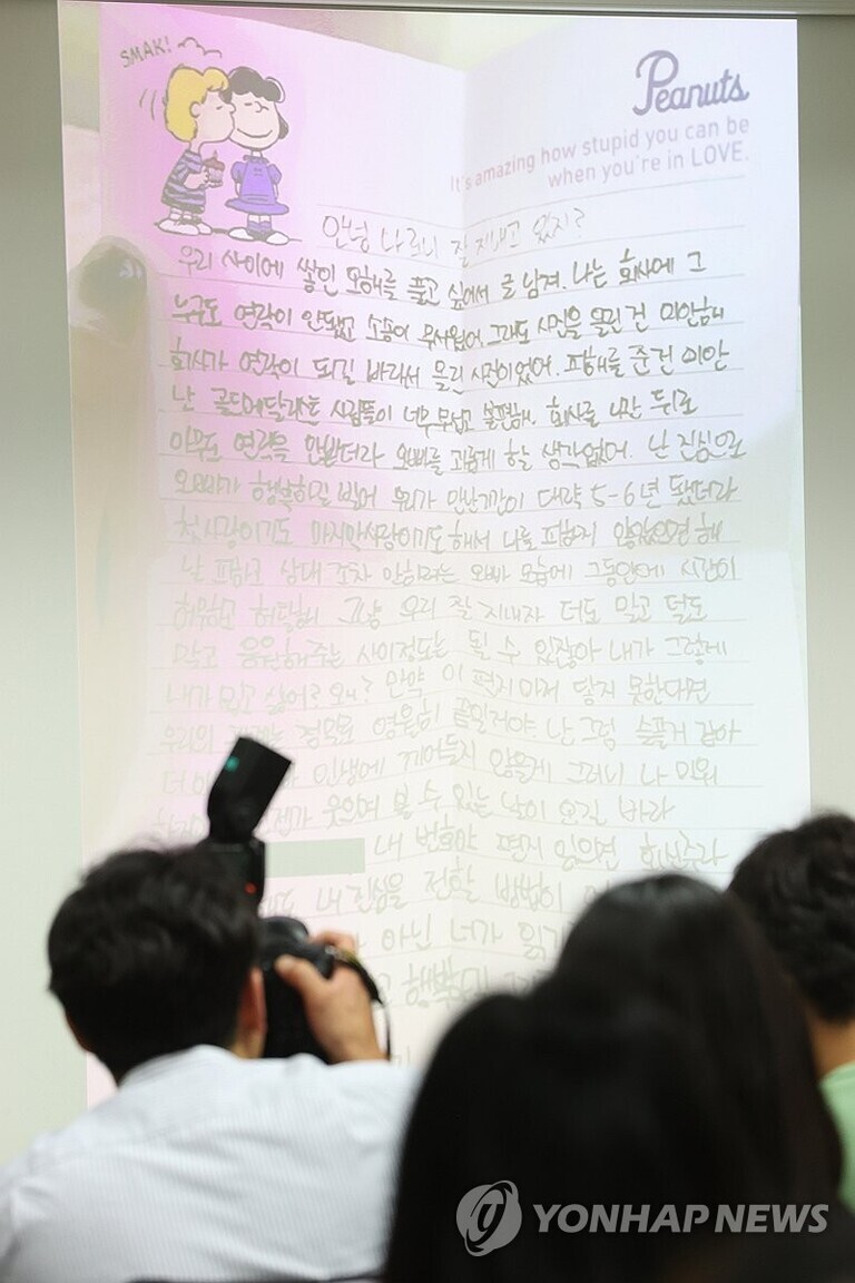 여러 명의 사람들이 앉아 있고, 앞에는 스누피 만화 캐릭터 그림과 영어 및 한글로 작성된 손편지가 화면에 크게 비춰져 있다. 편지 상단에는 'Peanuts'와 함께 두 사람이 포옹하는 일러스트가 보인다.