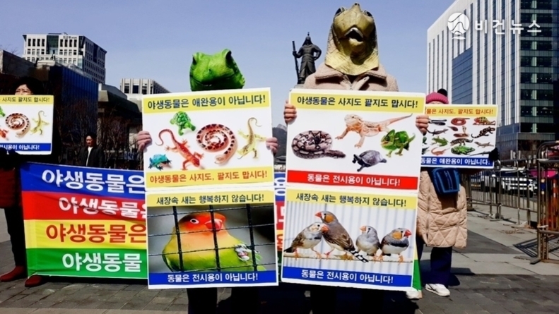 도심 거리에서 동물 머리 탈을 쓴 사람들이 야생동물 판매 및 전시 반대 피켓을 들고 시위하는 모습. 다양한 파충류와 새 사진이 포스터에 인쇄되어 있으며, '야생동물은 애완용이 아닙니다', '사지도 팔지도 맙시다' 등의 문구가 보임.