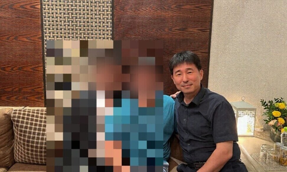 한 남성이 소파에 앉아 미소를 짓고 있고, 옆에는 모자이크 처리된 사람들이 함께 있다. 테이블 위에 꽃과 조명이 장식되어 있다.