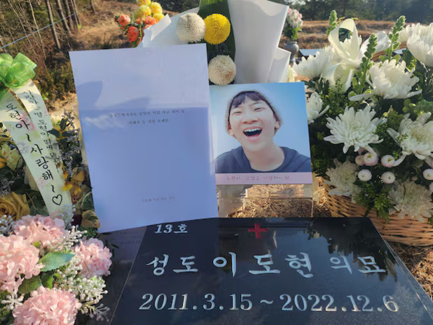 묘지에 꽃다발과 편지, 웃고 있는 젊은 사람의 사진이 놓여 있다. 묘비에는 '성도 이도현 의묘', 2011.3.15~2022.12.6이라는 글씨가 새겨져 있으며 주변에는 다양한 색상의 국화와 리본 장식이 보인다.