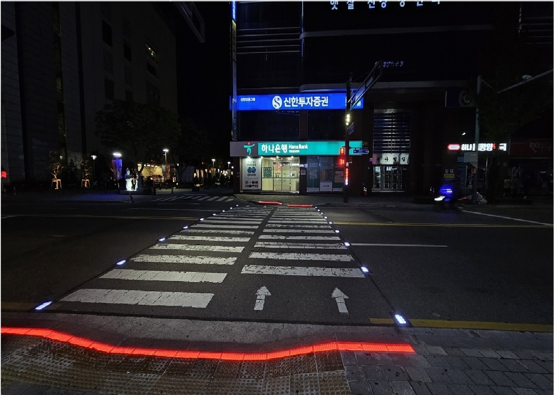 야간에 신호등 없는 횡단보도가 보이며, 횡단보도 양옆에는 파란색 LED 조명이, 인도 경계석에는 빨간색 LED 조명이 켜져 있다. 맞은편 건물 1층엔 하나은행과 다양한 상점이 있다.