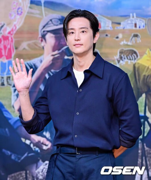 남성이 네이비색 셔츠와 바지를 입고 한 손을 들어 인사하는 모습. 배경에는 풍경과 사람, 동물이 그려진 포스터가 있다.