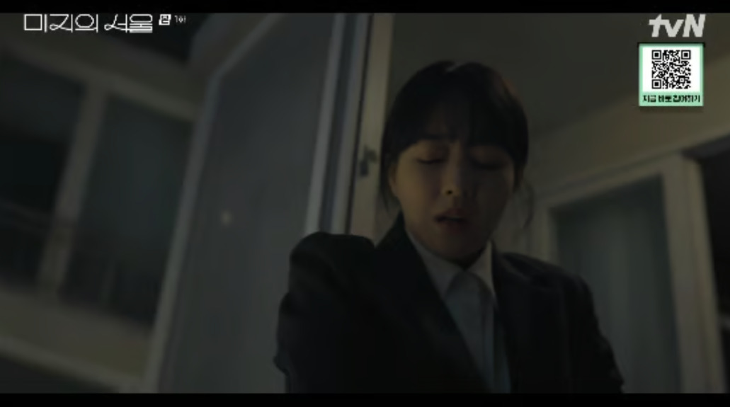 어두운 실내에서 정장을 입은 여성이 눈을 감고 고통스러운 표정을 짓고 있다. 배경에는 열린 문이 보인다.