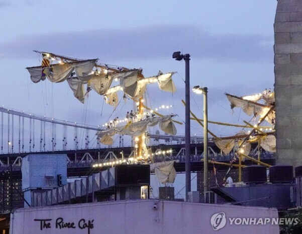 저녁 하늘 아래 조명이 켜진 범선의 돛대와 돛이 보이고, 앞쪽에는 'The River Café'라는 간판과 건물이 있다. 배경에는 다리 구조물도 보인다.