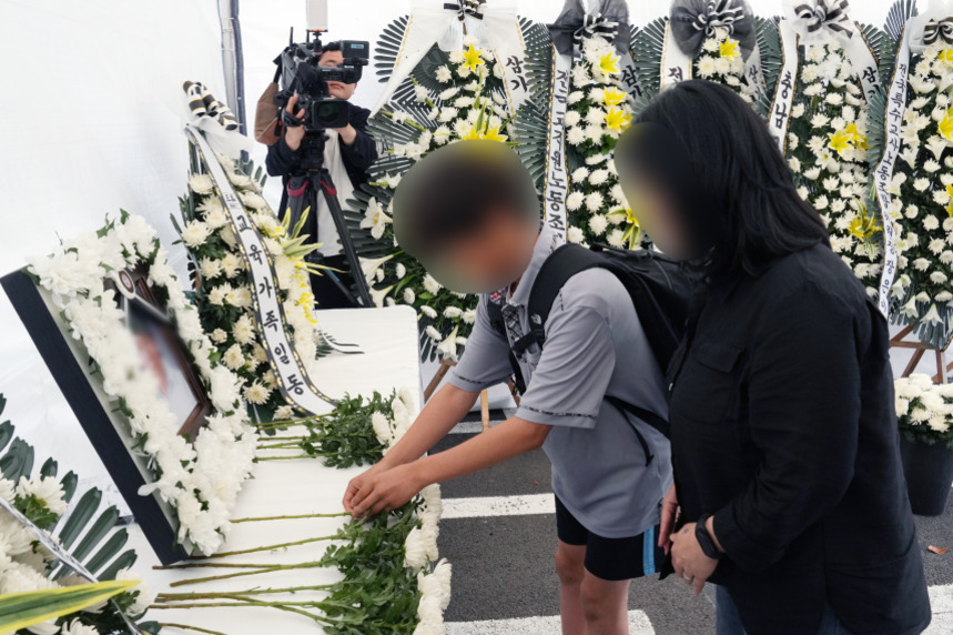 조문객들이 영정 사진 앞에 국화꽃을 올리며 헌화하고 있고, 뒤에는 근조 화환이 여러 개 놓여 있다.