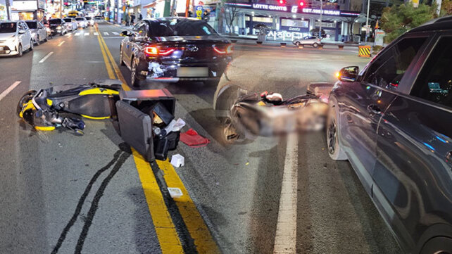 도심 도로 한복판에서 오토바이와 자동차가 충돌하여 오토바이가 쓰러져 있고, 일부 물건들이 흩어져 있는 교통사고 현장