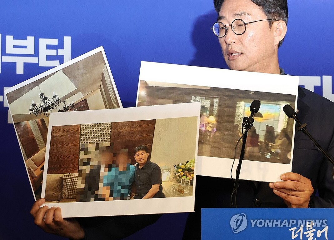 안경을 쓴 남성이 파란색 배경 앞에서 마이크 옆에 서서 여러 장의 사진이 인쇄된 종이를 들어 보이고 있다. 한 사진에는 모자이크 처리된 사람들이 함께 앉아 있고, 다른 사진들에는 실내 모습과 조명이 담겨 있다.