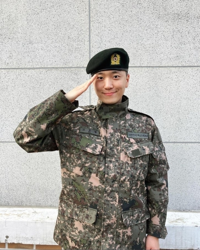 군복을 입고 베레모를 쓴 사람이 벽 앞에서 거수경례를 하고 있다.