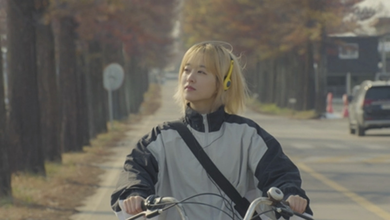 노란색 헤드폰을 쓴 단발 금발의 사람이 가로수길에서 자전거를 타고 있다.