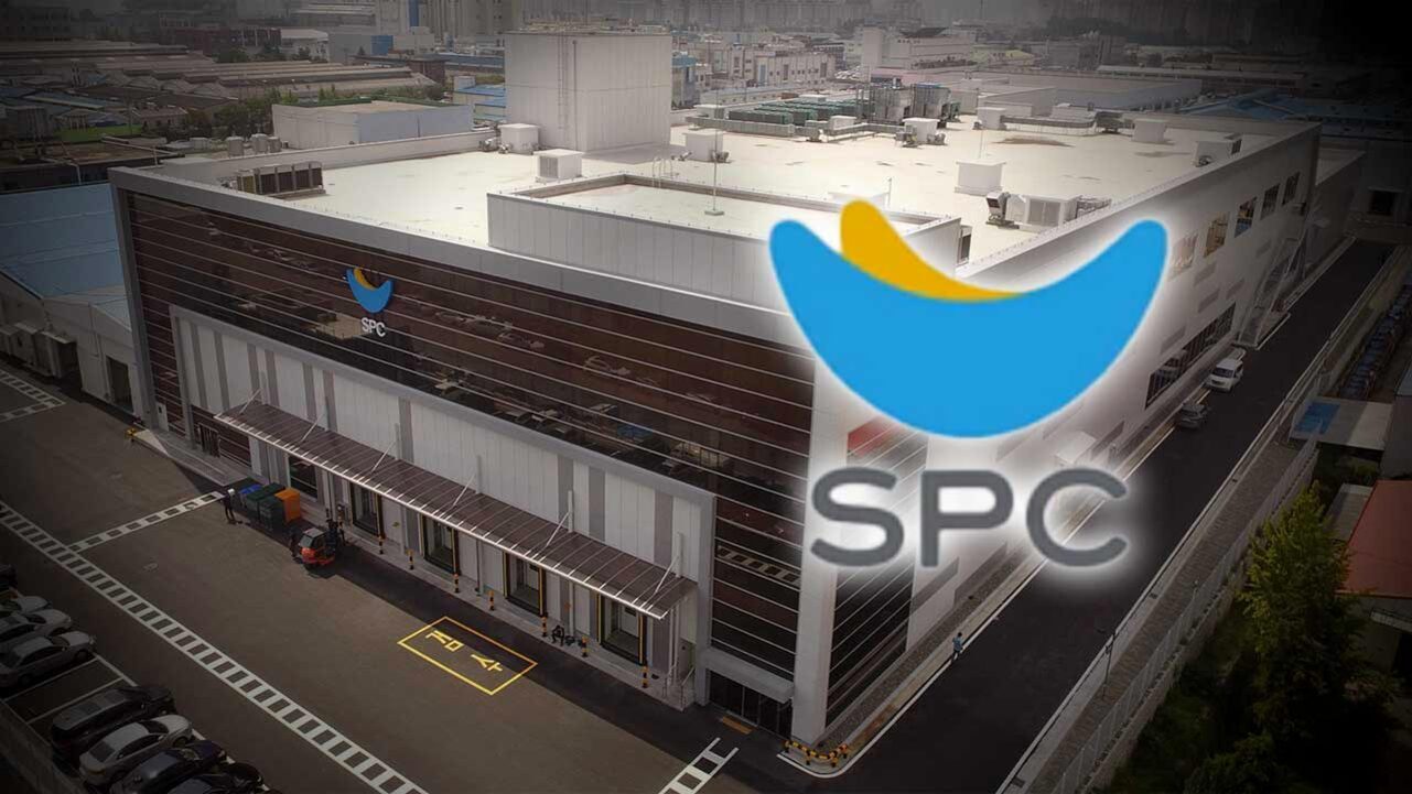 SPC 로고가 크게 보이고, 그 뒤로 대형 공장 건물과 주차장이 있는 산업 단지 전경이 나타나 있다.