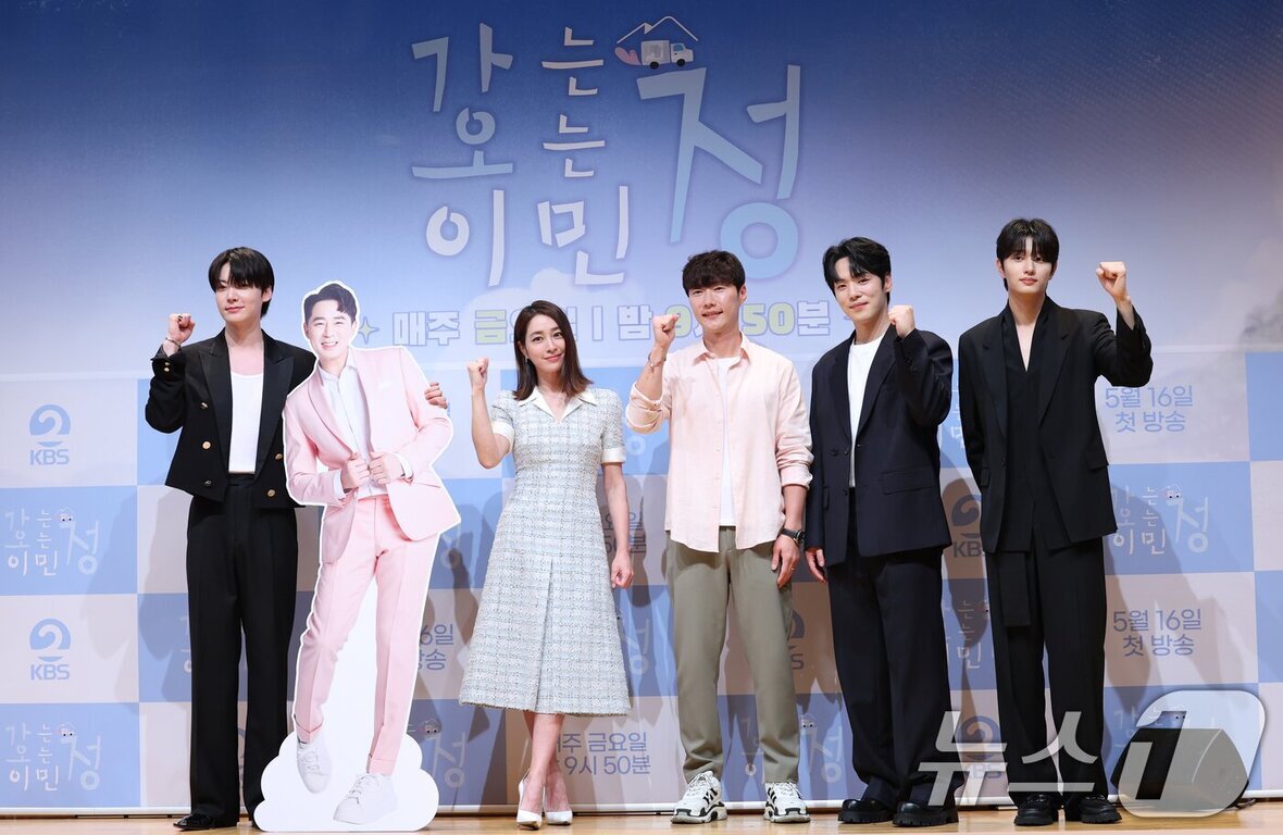 다섯 명의 사람들이 포토월 앞에서 서서 손을 들어 파이팅 포즈를 하고 있으며, 한 사람 옆에는 분홍색 정장을 입은 인물의 전신 등신대가 있다. 배경엔 '같이 사는 이민정'이라는 글자가 보인다.