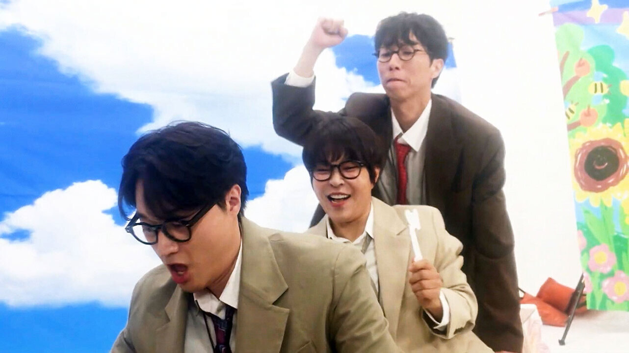 세 명의 남성이 교복과 안경을 착용하고 코믹한 포즈를 취하며 실내에서 촬영된 사진. 배경에는 파란 하늘 그림과 꽃이 그려진 판넬이 보인다.
