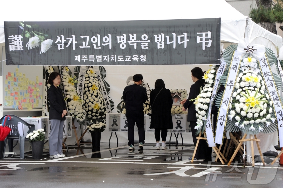 검은색 근조 현수막 아래 조문객들이 헌화하며 고인을 추모하는 야외 분향소 모습, 주변에는 하얀 국화꽃과 근조 화환이 놓여 있다.