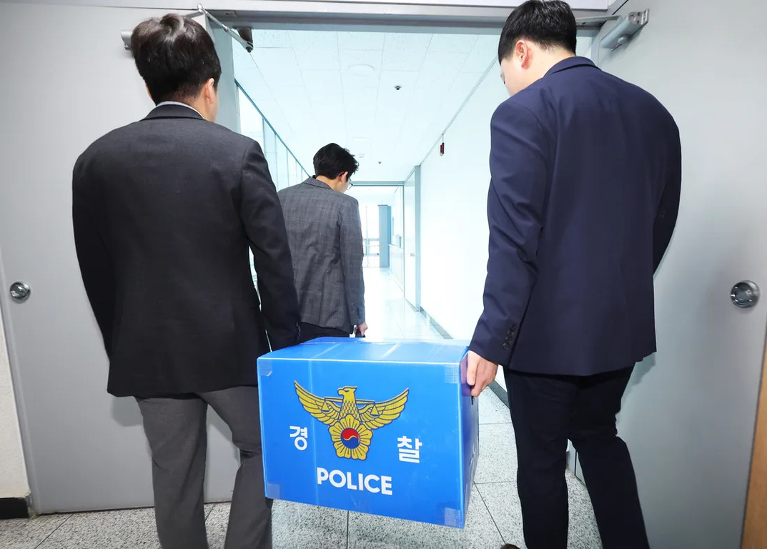 정장을 입은 남성 세 명이 경찰 로고와 '경찰 POLICE'가 적힌 파란색 상자를 들고 복도를 걷는 모습