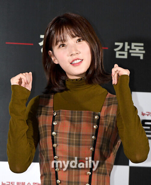 갈색 체크 원피스와 카키색 터틀넥을 입은 젊은 여성이 검정과 흰 배경 앞에서 두 손을 올리고 포즈를 취하고 있다.