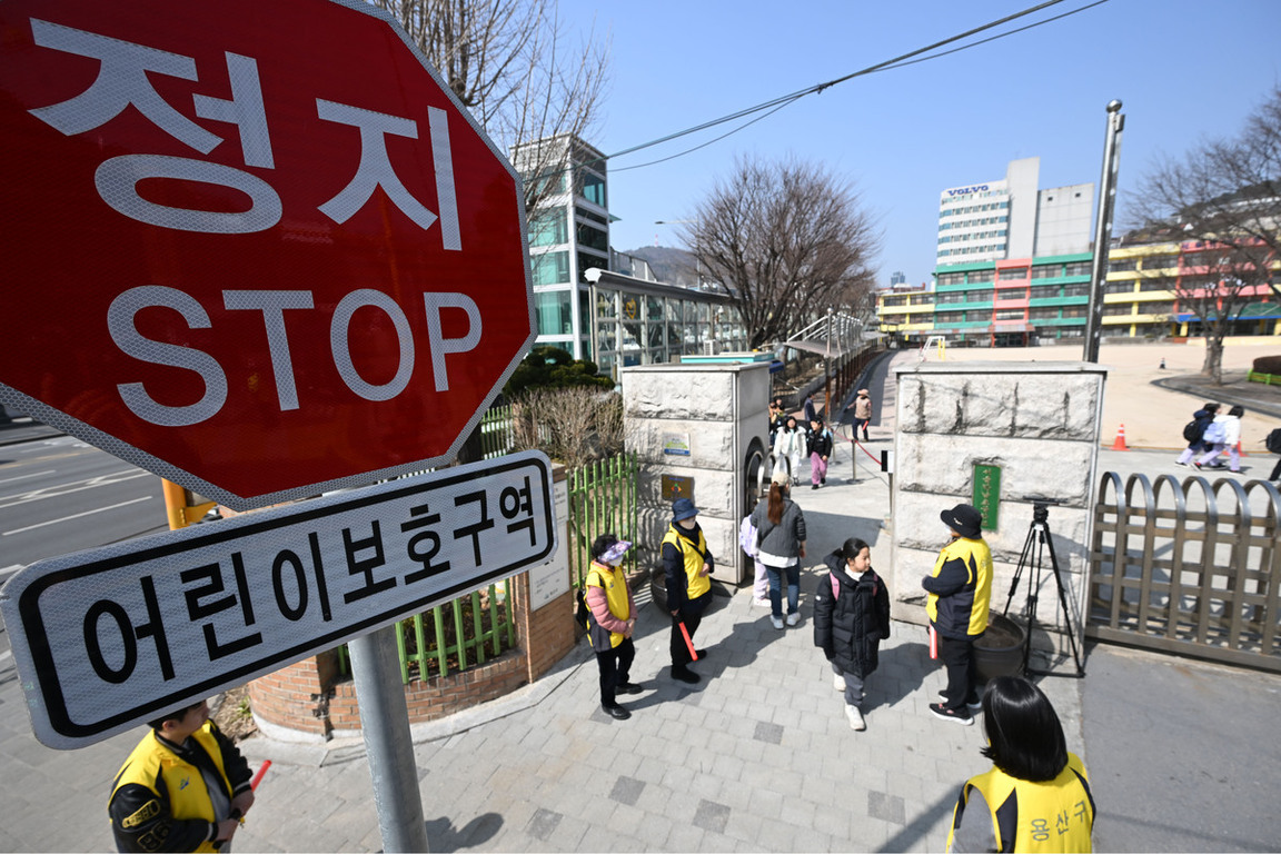 정지(STOP) 표지판과 어린이보호구역 안내문 아래로 학교 정문에서 등교하는 학생들과 노란 조끼를 입은 교통안전요원들이 보이는 모습
