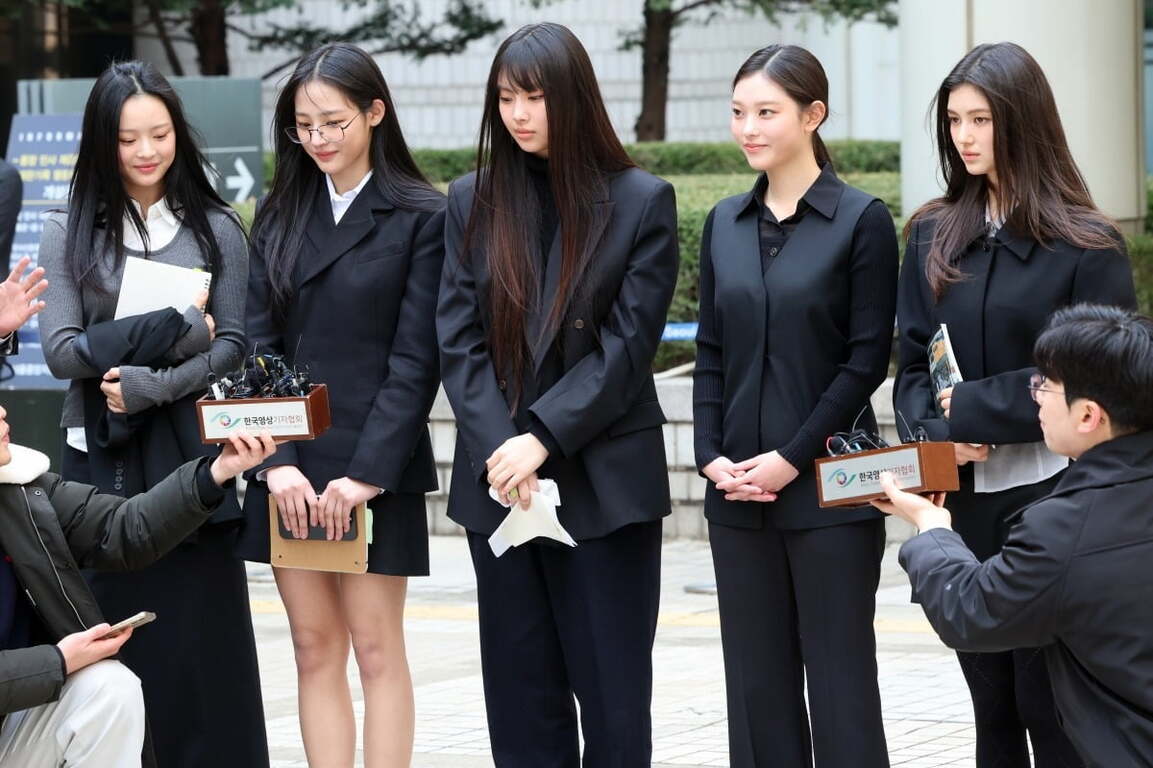 다섯 명의 젊은 여성이 정장 차림으로 바깥에서 기자회견을 하고 있으며, 앞에는 두 명의 남성 기자가 마이크를 들고 있다.
