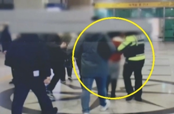 여러 명의 사람들이 실내 공간에서 모여 있고, 한 경찰관이 노란 원 안에 표시된 사람을 붙잡고 있다. 인물들의 얼굴은 흐리게 처리되어 있다.