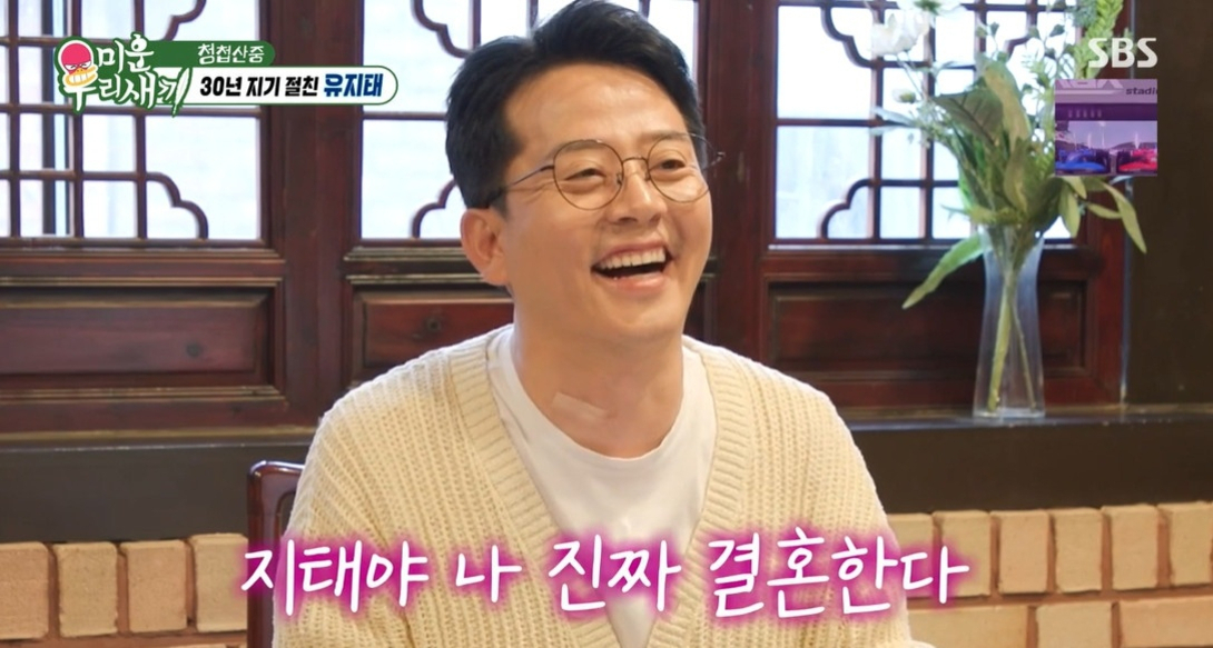 안경을 쓴 남성이 미소를 지으며 앉아 있고, 화면 하단에는 '지태야 나 진짜 결혼한다'라는 자막이 있다. 배경은 전통적인 창문과 꽃병이다.
