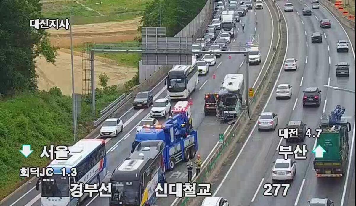 고속도로에서 버스와 차량의 교통사고 현장으로, 사고 처리 중인 견인차와 구급차가 있고 주변에 경찰과 도로 작업자가 보임. 일부 차선이 통제되어 정체가 발생하고 있음.