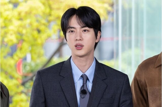검은 머리를 한 남성이 회색 정장을 입고 마이크를 단 채 실내에서 서 있는 모습
