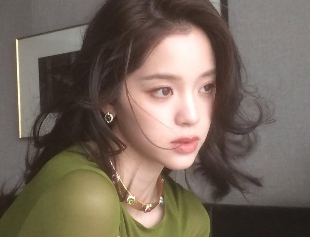 긴 흑발을 가진 여성이 초록색 상의를 입고 창밖을 응시하고 있다. 자연스러운 조명과 약간 흐트러진 머리카락이 특징이다.