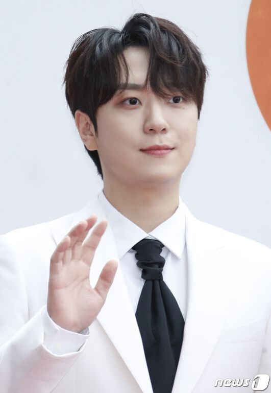 흰색 정장을 입고 검은 넥타이를 맨 남성이 한 손을 들어 인사하는 모습