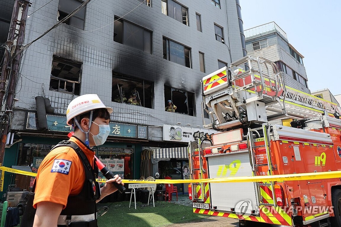 불이 난 건물 앞에 119 소방차와 구조대원이 있으며, 창문에는 그을음 자국과 함께 내부에서 작업 중인 소방관들이 보인다. 현장 주변은 통제선으로 차단되어 있다.