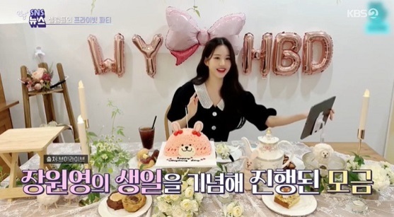 여성이 생일 파티 테이블 앞에 앉아 있고, 배경에는 'WY HBD' 풍선 장식과 큰 분홍색 리본이 있다. 테이블 위에는 케이크와 여러 음식들이 놓여 있으며, 화면 하단에 '장원영의 생일을 기념해 진행된 모금'이라는 자막이 보인다.
