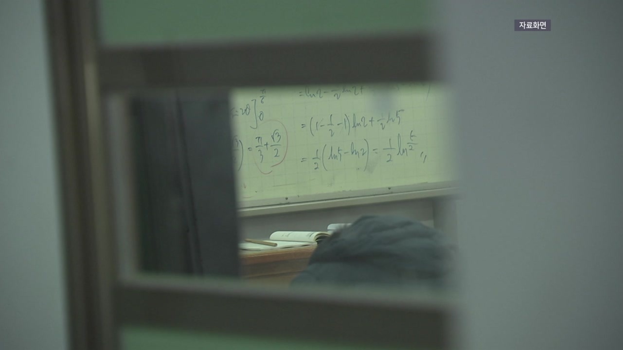 창문 너머로 본 교실 안에 수학 공식이 적힌 칠판과 책상 위에 펼쳐진 책, 머리를 숙이고 있는 사람이 보인다.