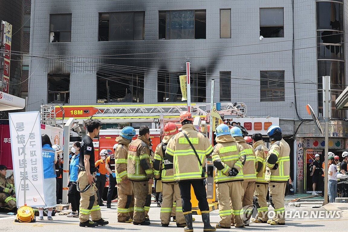불이 난 건물 앞에서 소방관들이 모여 상황을 점검하고 있으며, 창문과 벽에는 화재로 인한 그을음 자국이 남아 있다. 주변에 구조대원들과 응급 요원들도 보인다.
