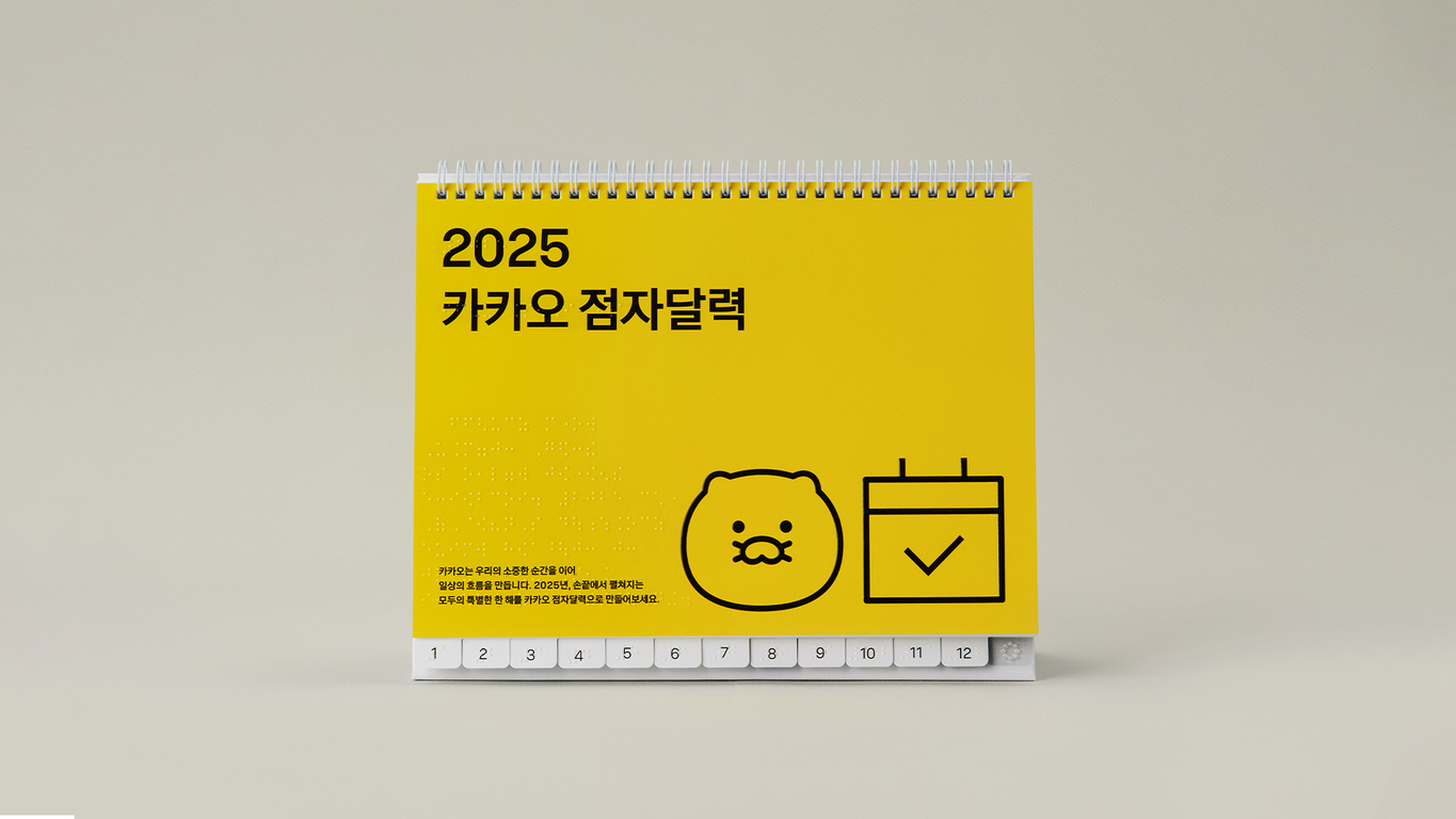 2025 카카오 점자달력이 쓰인 노란색 탁상달력 표지. 달력에는 귀여운 캐릭터 얼굴과 체크 표시가 있는 달력 아이콘이 그려져 있다. 하단에 1부터 12까지 월을 나타내는 흰색 버튼이 있고, 왼쪽에는 점자가 적혀 있다.