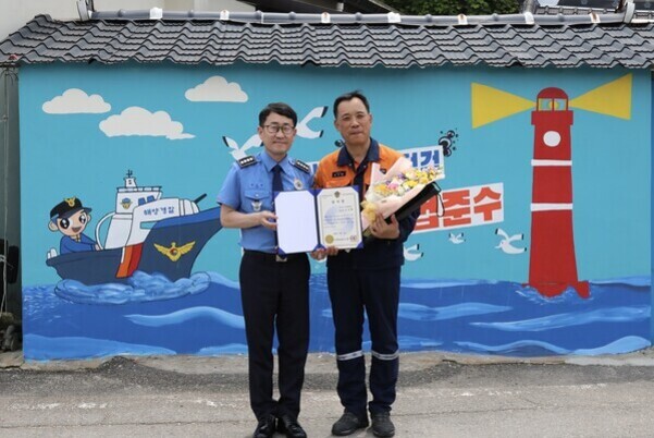 해양경찰 제복을 입은 남성과 소방관 제복을 입은 남성이 벽화 앞에서 상장과 꽃다발을 들고 기념 촬영하고 있다. 벽화에는 배와 등대, 갈매기가 그려져 있다.