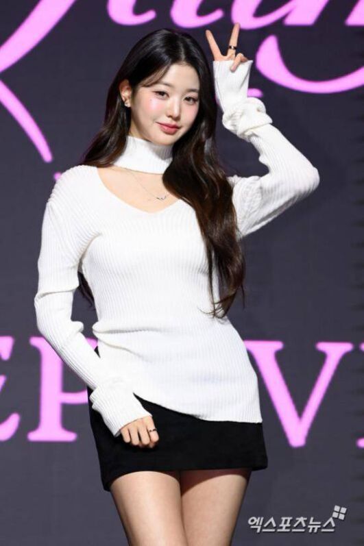 흰색 니트와 검은 치마를 입은 여성이 무대 앞에서 브이 포즈를 하고 있다.