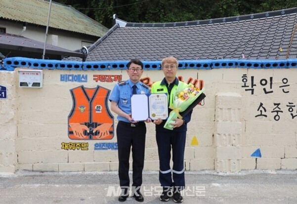 파란색 경찰 제복을 입은 남성과 노란색 조끼를 입은 남성이 벽 앞에 서서 상장과 꽃다발을 들고 기념사진을 찍고 있다. 벽에는 구명조끼 그림과 안전 문구가 그려져 있다.