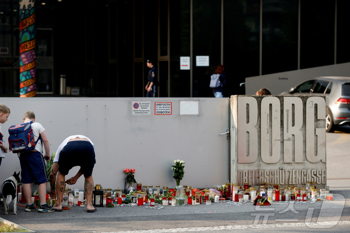 사람들이 BORG Dreierschützengasse라는 간판 앞에 촛불과 꽃을 두고 추모하고 있다. 벽 뒤에는 학교 건물로 보이는 곳이 있으며, 몇 명의 사람들이 지나가고 있다.