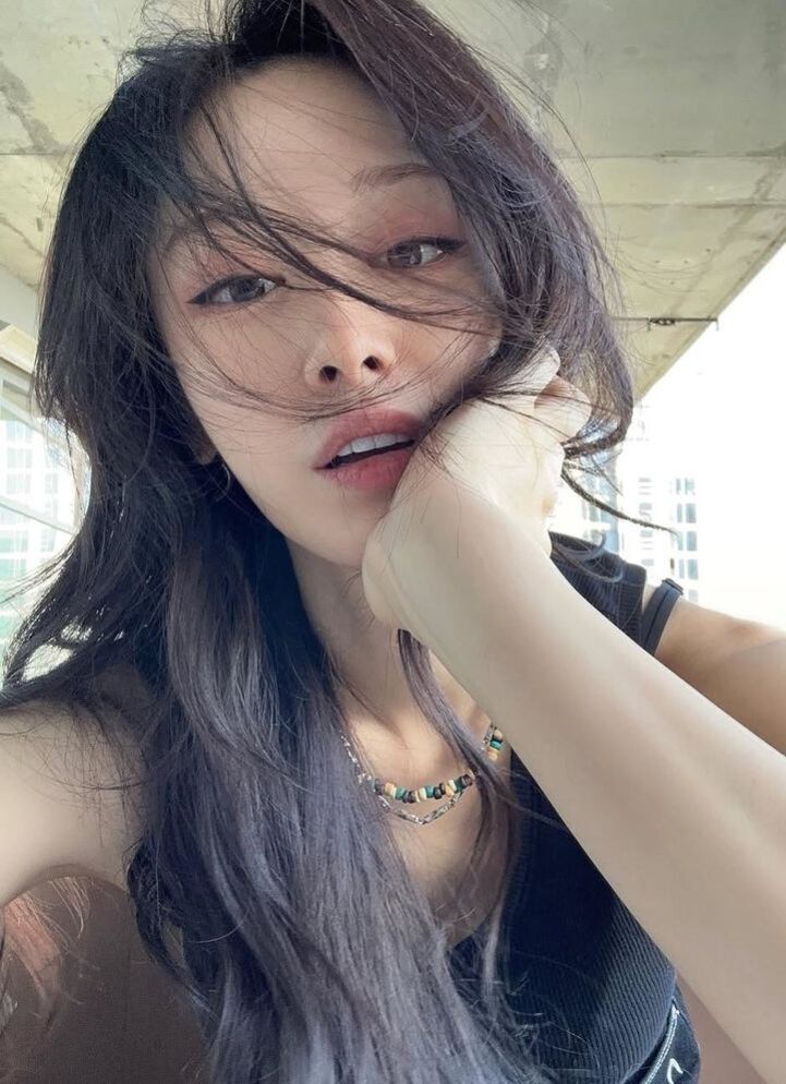 긴 머리를 가진 여성이 턱을 괴고 카메라를 응시하며 셀카를 찍는 모습. 바람에 머리카락이 얼굴 일부를 가리고 있다.