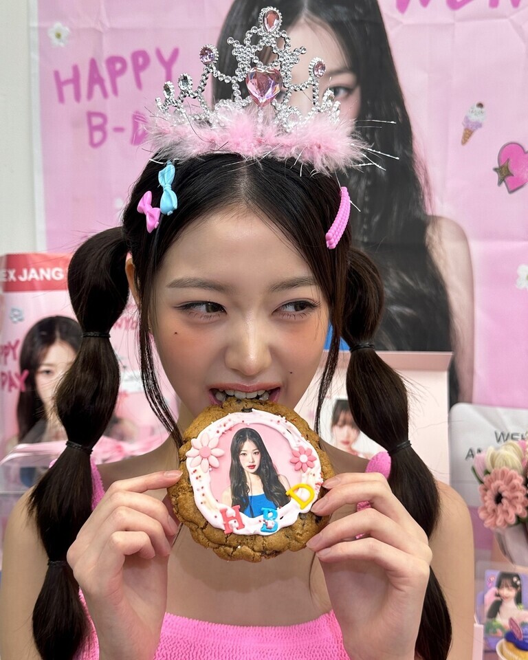 왕관을 쓴 젊은 여성이 양갈래 머리를 하고 HBD 글씨와 자신의 사진이 장식된 쿠키를 먹으려는 모습. 배경에는 분홍색 생일 축하 현수막과 해당 여성의 사진들이 보인다.