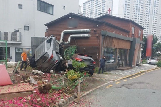 자동차 두 대가 건물 옆 화단에 충돌해 한 대는 기울어진 상태로 멈춰 있고, 주변에는 파손된 꽃과 구조물이 보이며 사람들이 현장을 살펴보고 있는 모습