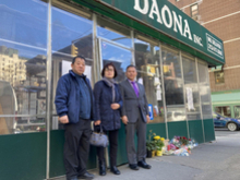 세 명의 성인이 DAONA INC라고 쓰인 가게 앞 인도에 서 있고, 오른쪽에는 꽃다발이 놓여 있다.