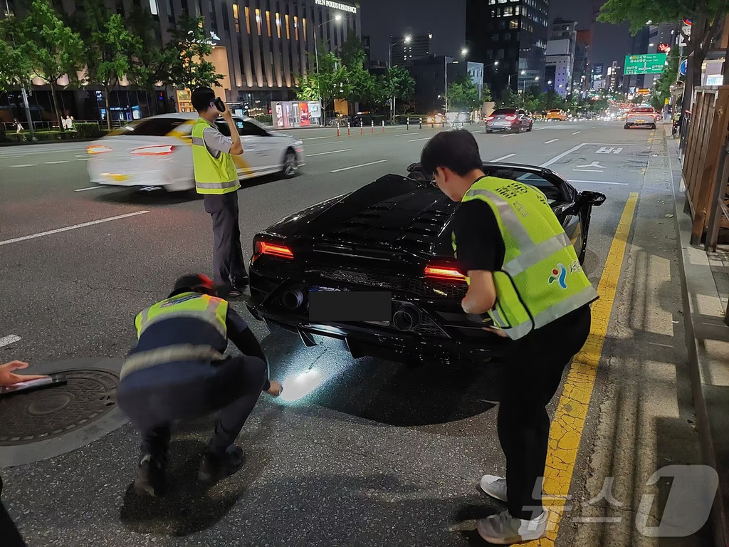 야간 도로에서 경찰 조끼를 입은 사람들이 고급 스포츠카의 배기구 근처를 손전등으로 조사하고 있는 모습
