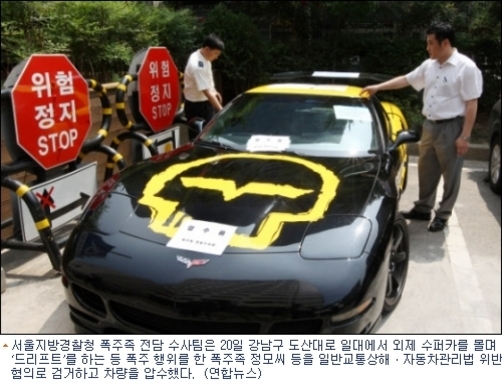 검은색 스포츠카 보닛에 노란 박쥐 로고가 크게 그려져 있으며, 차량 위에는 압수 관련 문서들이 놓여 있다. 두 남성이 차 옆에서 상황을 확인 중이고, 뒤편에는 '위험 정지' 표지판이 설치되어 있다.