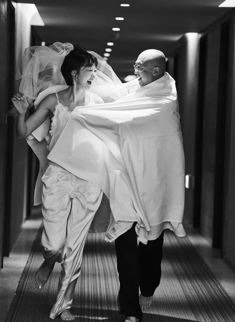 복도를 달리며 서로를 바라보며 웃고 있는 신부와 머리에 이불을 두른 남성의 흑백 사진
