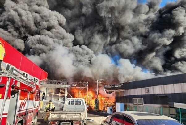 대형 화재가 발생한 건물에서 검은 연기가 치솟고 있으며, 소방차와 소방관이 출동해 진압 작업을 하고 있다.