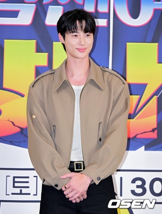 짧은 검정 머리의 남성이 베이지색 재킷과 흰 티셔츠를 입고 포스터 앞에서 미소 짓고 있다.