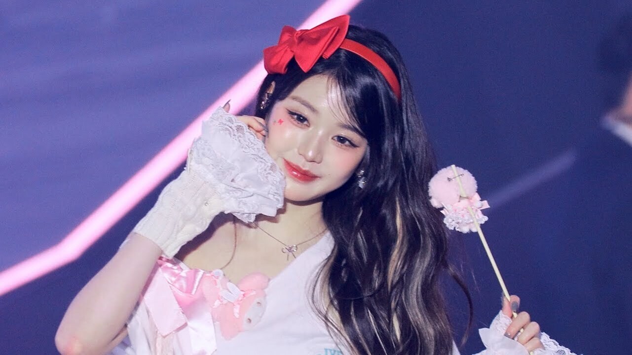 붉은 리본 머리띠와 레이스 장갑, 분홍색 소품을 든 긴 머리의 여성이 무대 위에서 포즈를 취하고 있다.