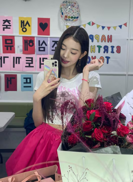 하얀 티셔츠와 분홍색 치마를 입은 젊은 여성이 거울 셀카를 찍고 있다. 앞에는 빨간 장미꽃다발과 선물 상자가 놓여 있고, 뒤 배경에는 생일 축하 문구가 붙어 있는 벽이 보인다.