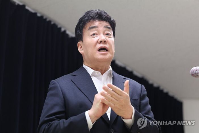 정장 차림의 남성이 마이크 앞에서 손짓을 하며 말하고 있다.