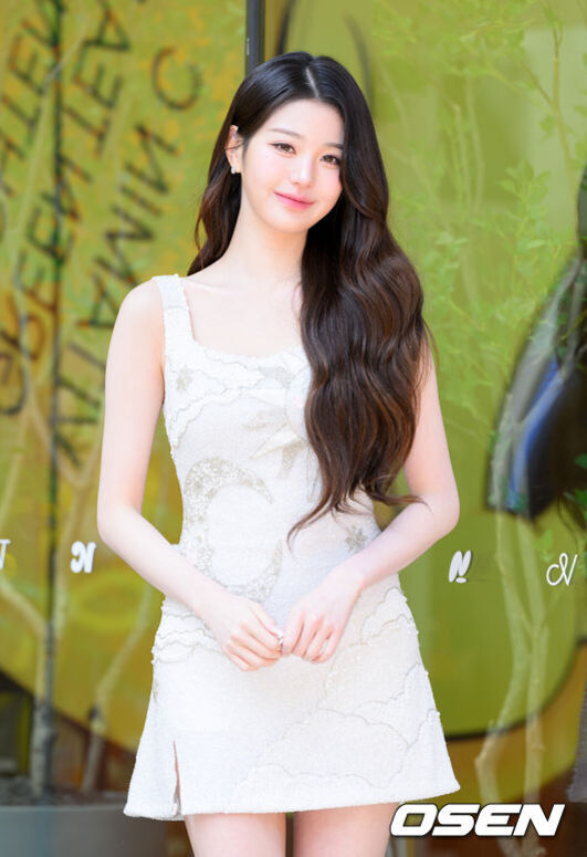 긴 웨이브 머리를 한 여성이 흰색 민소매 원피스를 입고 야외에서 미소를 짓고 있는 모습